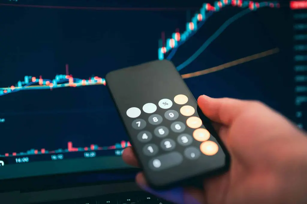 Hand using smartphone calculator over laptop displaying financial stock market trading charts