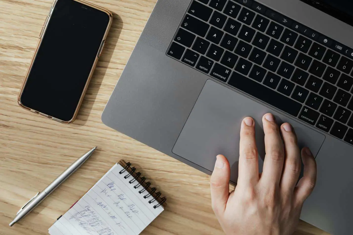 Hand on laptop, smartphone, notebook, pen on a light wooden desk workspace