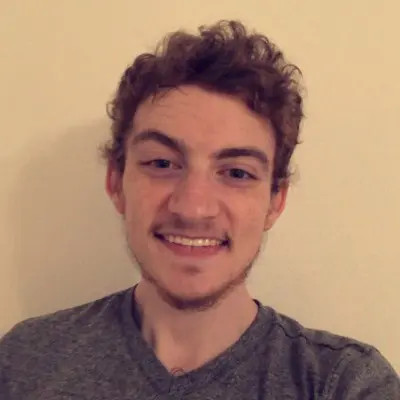 Smiling young man with curly brown hair, freckles, and a light beard