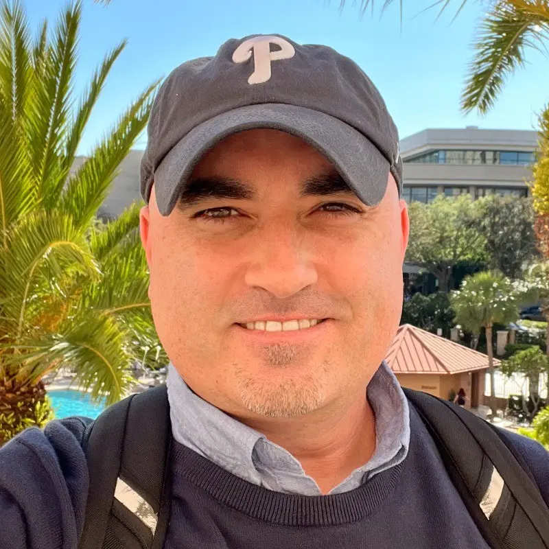 Man smiling wearing Phillies baseball cap outdoors with palm trees and building