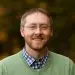 Smiling man with glasses and beard wearing a green sweater and plaid shirt